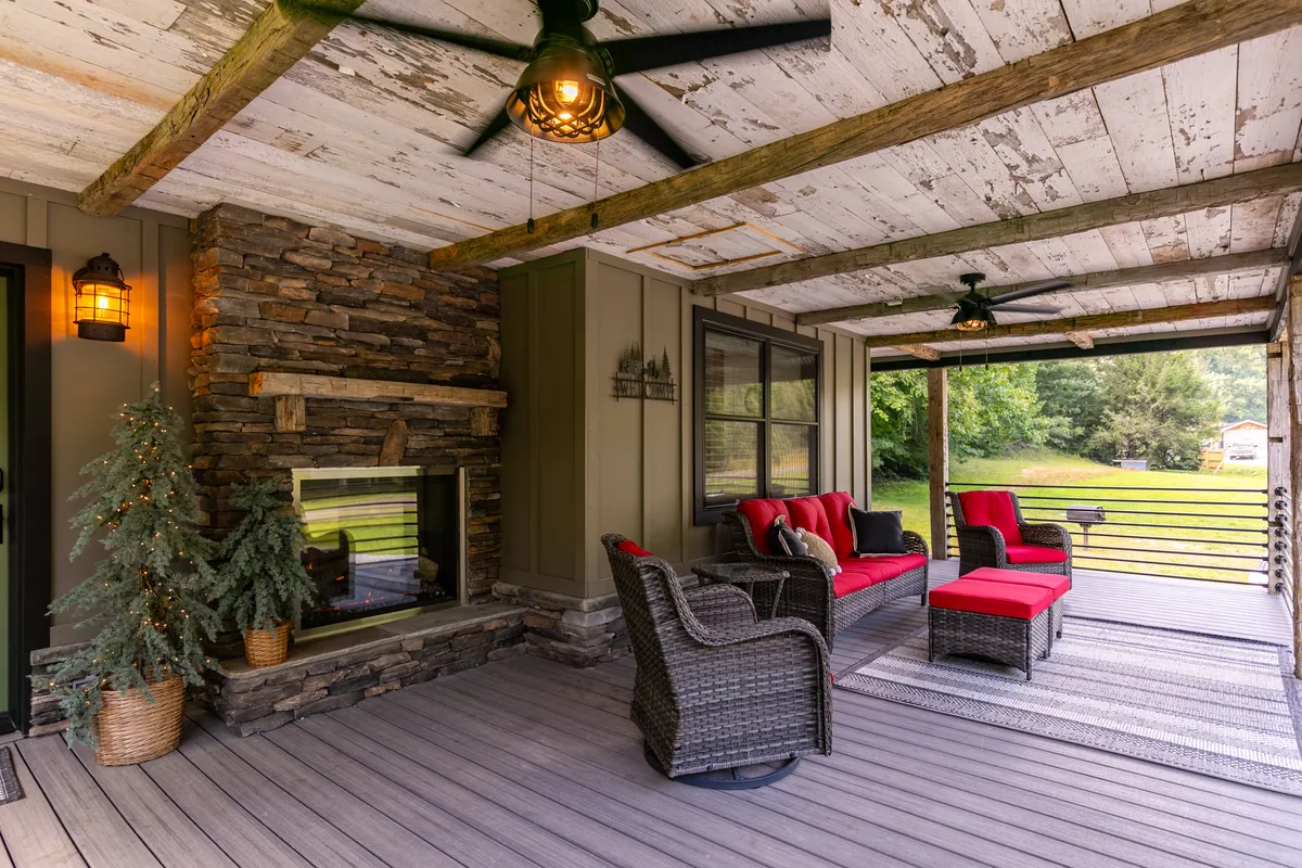 The image captures a portion of the wrap-around covered deck, featuring a prominent stone outdoor fireplace, plush wicker furniture with red cushions, and ceiling fans overhead. Tall trees frame the background, blending the comfort of the deck with the surrounding nature. The atmosphere feels cozy and welcoming, perfect for relaxing or entertaining outdoors.