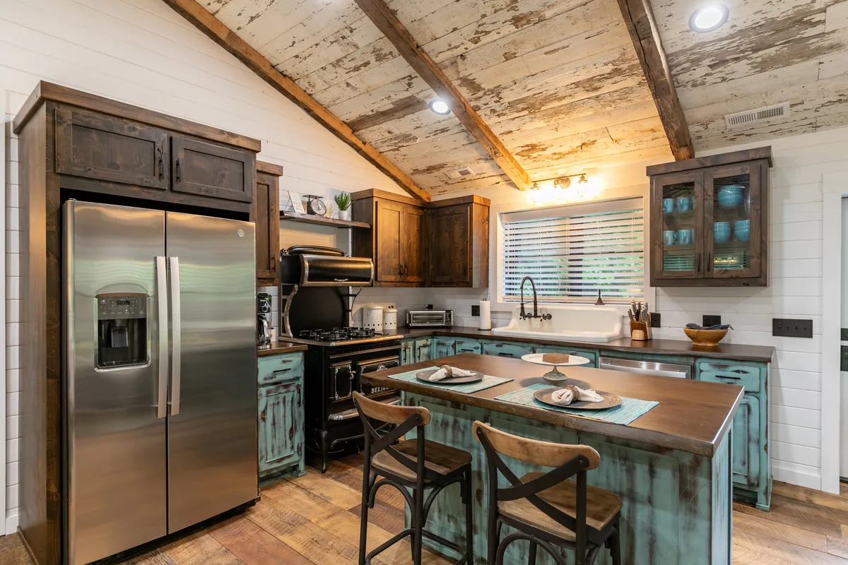 An open view of a kitchen and dining area with a rustic wooden ceiling, turquoise cabinets, stainless steel appliances, and a dining table set for a meal. Natural light fills the space, and the setup hints at shared family gatherings and cozy meals.