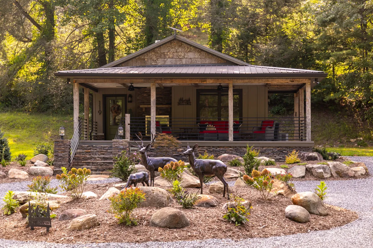 The image showcases the stone house with a broad front porch, deer sculptures, and a landscaped garden. The porch and garden provide an ideal setting for outdoor gatherings and connecting with nature, reinforcing the home's seamless indoor-outdoor flow.