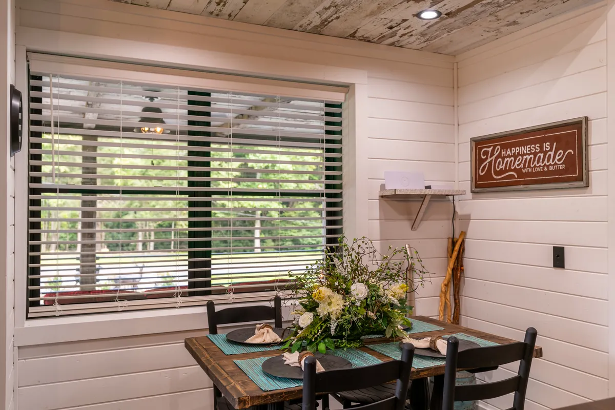 A welcoming dining table decorated with a floral centerpiece, placed next to windows with blinds that let in soft daylight. Thoughtful wall art contributes to the cozy atmosphere, evoking a sense of togetherness and homey comfort.