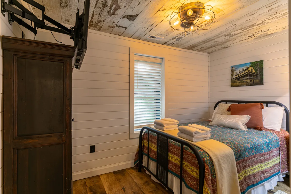 A welcoming bedroom highlighted by a bed with a colorful quilt, a wooden wardrobe, large window with blinds, and tasteful wall art. The space glows with natural light, emphasizing the room's coziness and suitability for peaceful slumber.