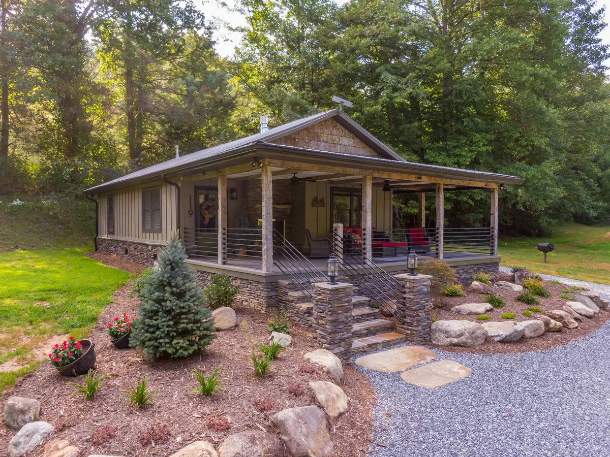 The image shows the inviting exterior of a modern farmhouse nestled amid lush greenery in Linville, North Carolina. Stone steps lead up to a charming porch with elegant railings, decorative lanterns, and glimpses of vibrant garden flowers. The tranquil backdrop hints at surrounding woods and a nearby creek, all bathed in the warm glow of sunset.