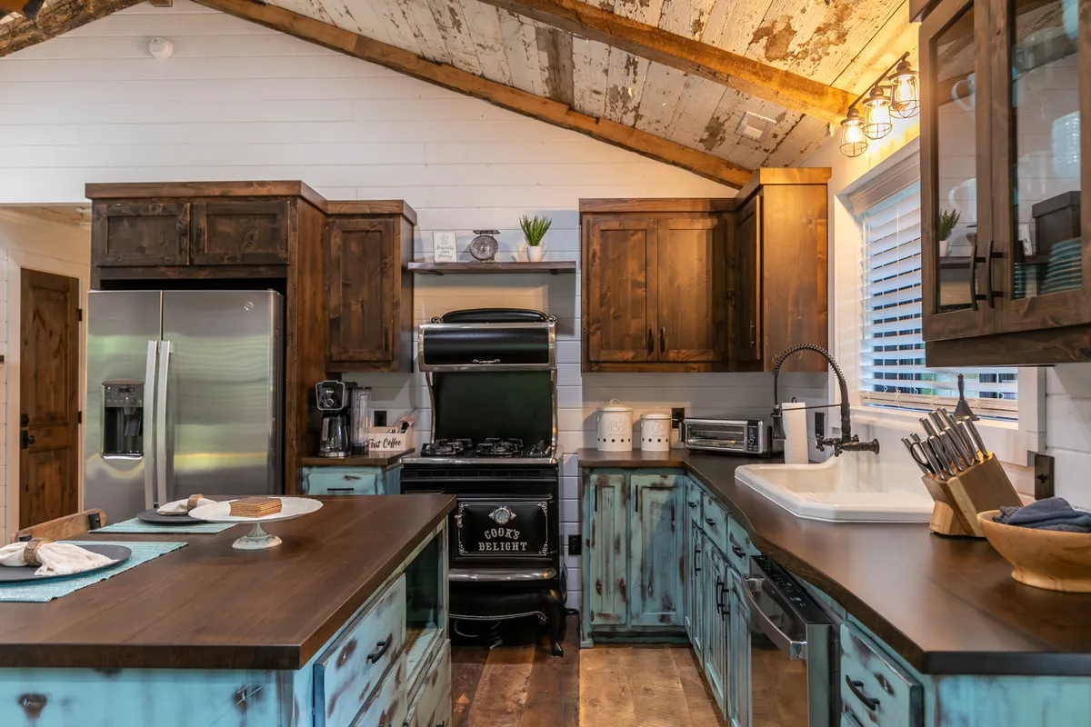 A bright, inviting kitchen featuring a central island with a dark wooden countertop, rustic cabinetry accented with teal, gleaming stainless steel appliances, and decorative items neatly arranged on the counters. The space radiates warmth and modern farmhouse charm, making it an ideal setting for preparing meals or entertaining.