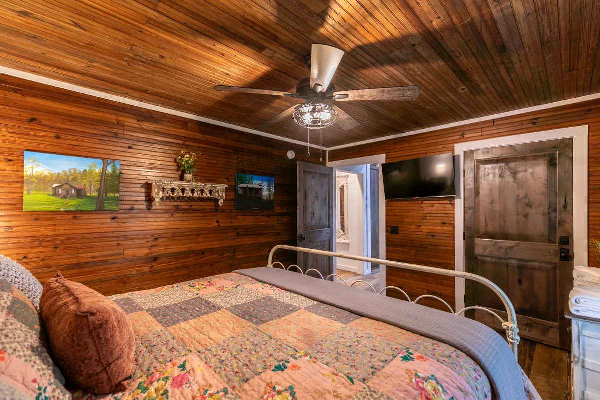 This image showcases a tranquil bedroom featuring a cozy bed with a quilt, warm wooden ceiling, wall art, and a ceiling fan. Soft natural light filters through the window, enhancing the inviting ambiance, while the ceiling fan and subtle decor create a comfortable environment for relaxation.