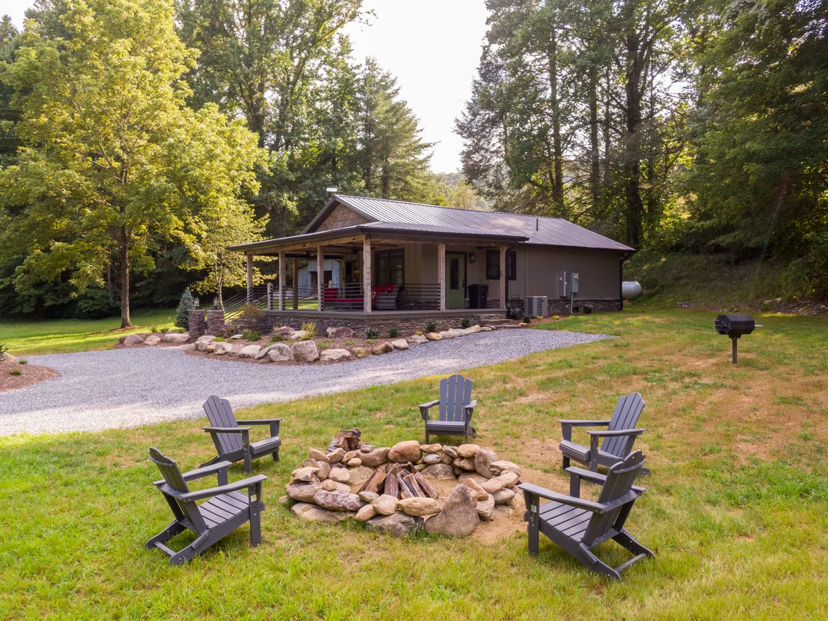 The photo centers on a fire pit area in the manicured yard, surrounded by Adirondack chairs and tall trees. The house’s exterior is visible in the background. The setting suggests an ideal gathering spot for evening conversations, s’mores, and stargazing under mountain skies.