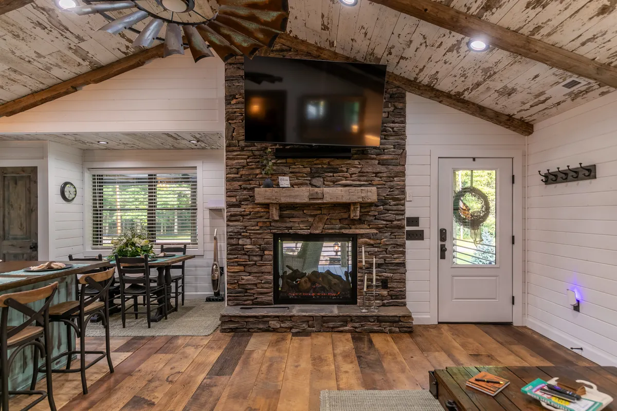 The image showcases a warm, inviting living room with a striking stone fireplace that acts as a focal point. Wooden beams add rustic charm overhead. A dining table with chairs sits nearby, and a television is mounted above the fireplace. The space looks spacious yet cozy, ideal for family gatherings or relaxing by the fire.