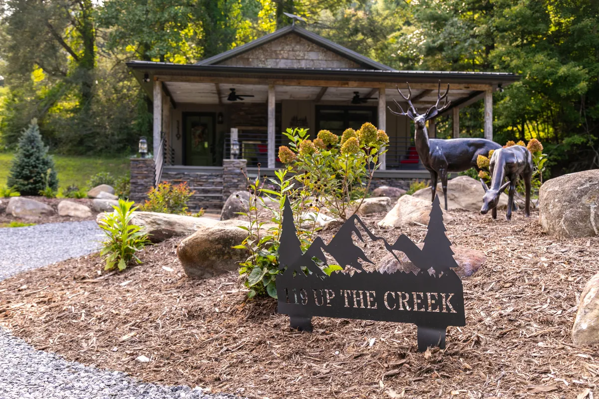This image features the exterior of the house, framed by mature landscaping and a charming deer sculpture. The welcoming porch hints at comfortable outdoor living and the landscaping sign adds a personalized touch. The serene setting showcases the home's semi-remote location and harmonious connection to nature.